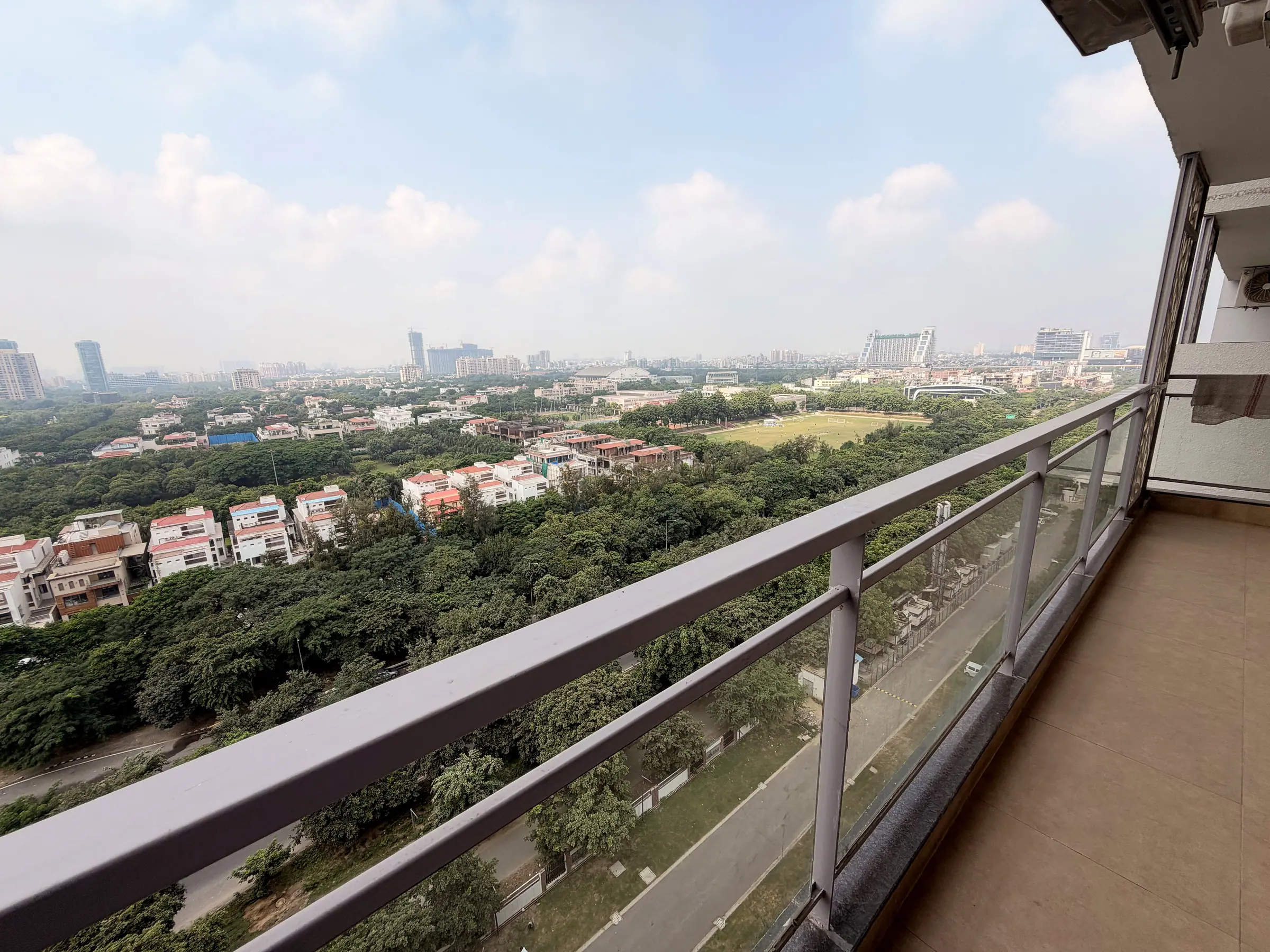 Balcony with a glass railing looking across the treetops, villas and greens to the Greater Noida skyline